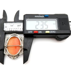 May include: An antique cameo brooch with an orange oval center, set in a silver-toned filigree frame. The brooch is being measured with a digital caliper, showing a measurement of 23.5 mm.