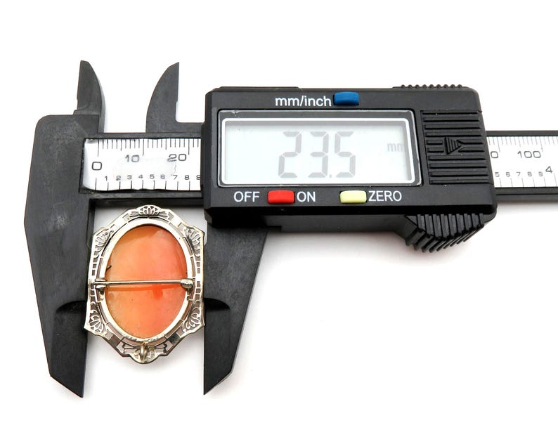 May include: An antique cameo brooch with an orange oval center, set in a silver-toned filigree frame. The brooch is being measured with a digital caliper, showing a measurement of 23.5 mm.