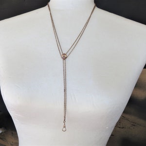 May include: A vintage necklace with a long, dangling chain. The necklace has a small, square pendant with pink gemstones and a clasp at the end. The necklace is displayed on a white mannequin.
