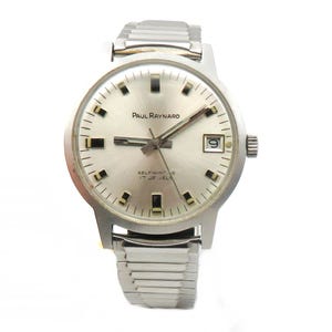 May include: A silver wristwatch with a silver band. The watch face is white with black markings and the text "PAUL RAYNARD" and "SELF WINDING 17 JEWELS".
