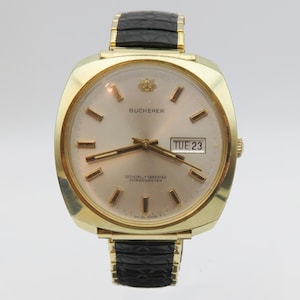 May include: A gold-toned wristwatch with a square face and a black leather band. The watch face has a white background with black hour markers and hands. The watch also has a day and date window at the 3 o'clock position. The watch face says "Bucherer" and "Officially Certified Chronometer".