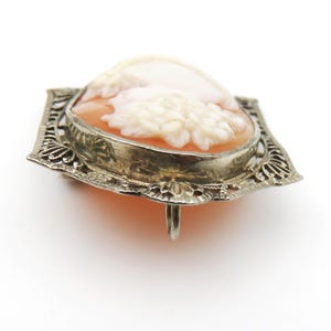 May include: A vintage cameo brooch featuring a carved profile set in a silver-toned filigree frame. The cameo has a peach-colored background with a white relief carving. The brooch is a classic piece of jewelry.