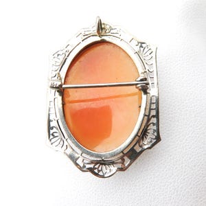 May include: A vintage cameo brooch with an oval, orange-toned center set in a silver-colored filigree frame. The brooch has a pin closure on the back and a small loop at the top for hanging.