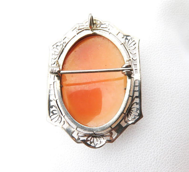 May include: A vintage cameo brooch with an oval, orange-toned center set in a silver-colored filigree frame. The brooch has a pin closure on the back and a small loop at the top for hanging.