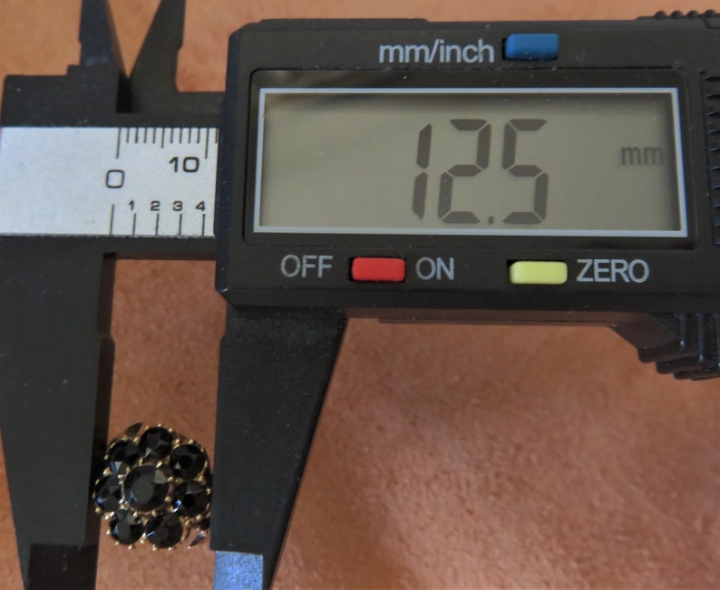 May include: A black and gold button with a diameter of 12.5 millimetres, measured with a digital caliper.