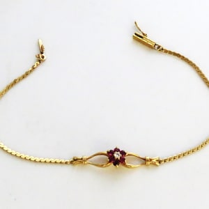 May include: A gold chain bracelet featuring a central floral motif composed of six deep red rubies surrounding a single clear gemstone. The bracelet includes a delicate link chain and a secure metal clasp mechanism at the ends.