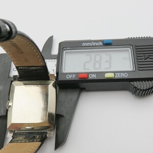 May include: A vintage rectangular wristwatch with a silver-colored face and black leather band. The watch is being measured with a digital caliper, showing a width of 28.3 mm (1.1 inches). The watch band is black with a crocodile pattern.