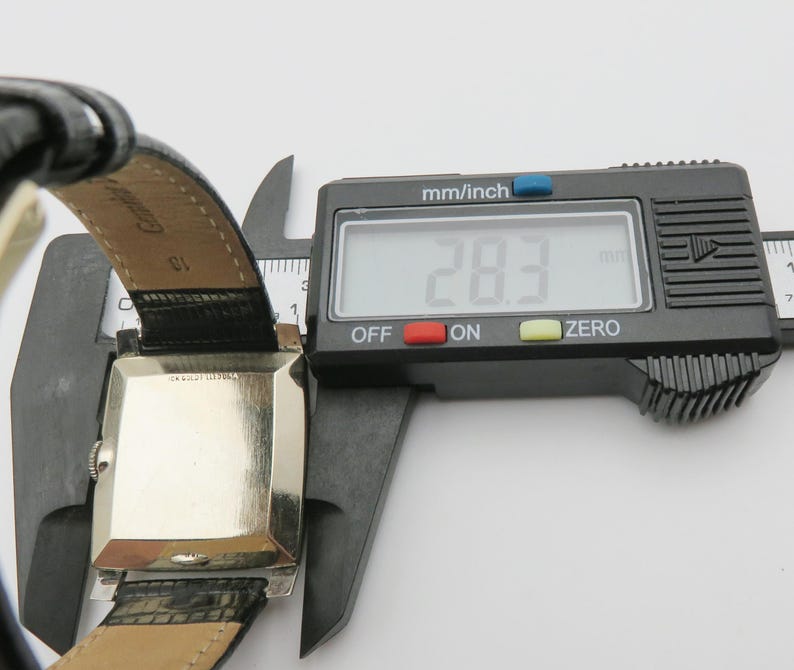 May include: A vintage rectangular wristwatch with a silver-colored face and black leather band. The watch is being measured with a digital caliper, showing a width of 28.3 mm (1.1 inches). The watch band is black with a crocodile pattern.