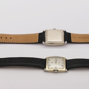 May include: Two vintage wristwatches with rectangular and square silver-toned cases and black leather bands. One watch has a light brown leather band on the inside. The watch faces have a light-colored background with black hands and markers.
