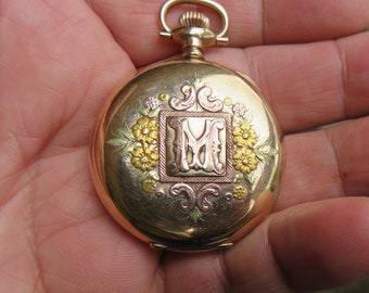 May include: A gold pocket watch with an engraved floral design and the letter "M" in the center.