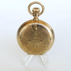 Puede incluir: Un reloj de bolsillo de oro con un diseño ornamentado en la parte posterior. El reloj tiene una esfera redonda y una tapa con bisagras. El reloj está en un soporte transparente.