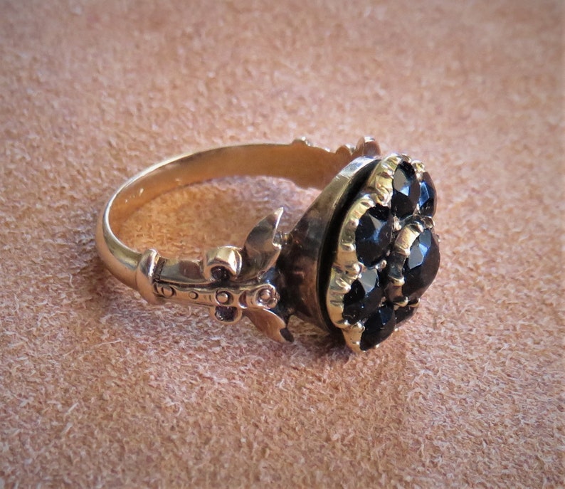 May include: A gold ring with a black stone cluster set in a bezel setting. The ring has a decorative band with a fleur-de-lis design.