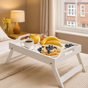 May include: A white wooden breakfast tray with folding legs holds a white ceramic coffee cup on a saucer, a yellow mug, a glass jar with a spoon, two yellow bananas, and a white plate topped with a waffle, blueberries, and banana slices.