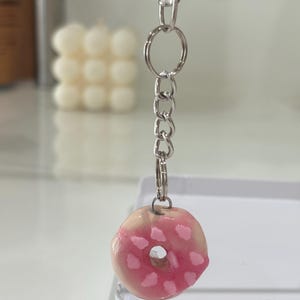 Peut inclure: Un porte-clés avec une breloque en forme de petit beignet circulaire, doté d'une base beige brillante, d'un glaçage rose et de petites décorations roses en forme de cœur. La breloque est fixée à une chaîne en métal argenté avec un anneau.