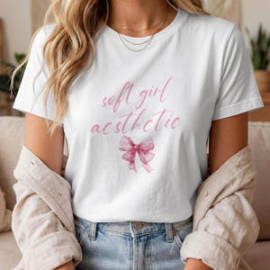 Könnte beinhalten: Ein weißes T-Shirt mit kurzen Ärmeln und dem in rosa Schreibschrift gedruckten Text soft girl aesthetic auf der Brustmitte. Unter dem Text befindet sich eine Illustration im Aquarellstil, die eine rosa Schleife darstellt.