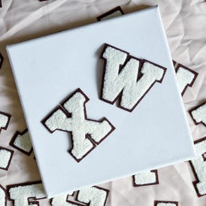 Op de afbeelding: Een verzameling witte letterpatches in varsity-stijl met donkerbruine randen, verspreid rond een centraal vierkant wit canvas. De patches hebben een getextureerd, pluizig oppervlak en zijn in een willekeurig patroon gerangschikt.