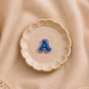 May include: A small, fabric-stitched letter A patch featuring a dark blue center with a light purple border. The patch is centered on a small, round, scalloped-edge ceramic dish. The letter is crafted with visible embroidery stitching along the edges.