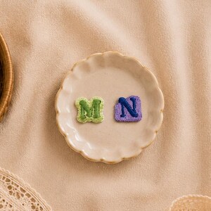 Puede incluir: Dos pequeños parches de tela bordados con las letras M y N en verde y morado respectivamente, sobre un plato de cerámica festoneado junto a un carrete de hilo de madera, tijeras de metal dorado y un borde de encaje beige.