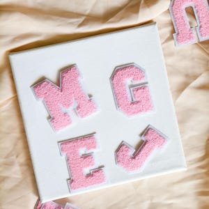 May include: A square white canvas featuring four pink chenille varsity-style letters arranged in a grid. The letters M, G, E, and J are attached to the surface. Additional loose pink chenille letters are visible at the top and bottom edges.