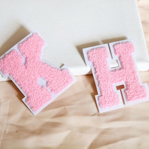 May include: Two varsity-style letter patches, the letters K and H, featuring a textured, fuzzy pink chenille surface with a white felt border. The letters are block-shaped with a classic collegiate font design, showcasing a soft, raised pile.