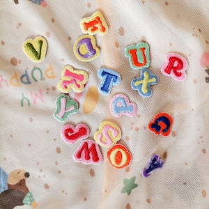 May include: A collection of small, colorful embroidered alphabet letter patches scattered on a light-colored mesh fabric featuring a repeating pattern of brown cartoon animals and small brown dots. The patches are in various bright colors.