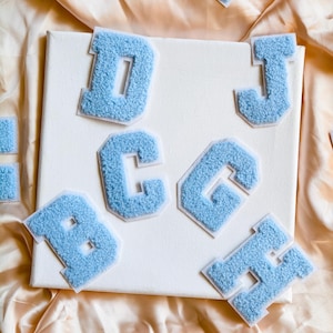 Peut inclure: Une collection de lettres thermocollantes bleu clair de style universitaire avec une surface en chenille texturée et duveteuse et une bordure blanche. Les lettres D, J, C, G, B et H sont disposées sur une toile carrée blanche.