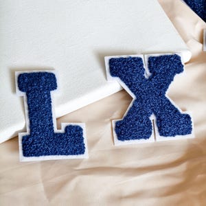 Op de afbeelding: Twee marineblauwe letterpatches in varsity-stijl, de letters L en X, met een getextureerd, pluizig chenille-oppervlak. Elke letter is omlijnd met een witte vilten rand, wat zorgt voor een duidelijk contrast met de diepblauwe stof.