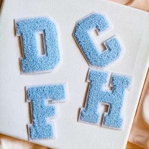 May include: Four light blue varsity-style letter patches featuring a textured, fuzzy chenille surface. The letters D, C, F, and H are arranged on a white surface, each outlined with a thin, clear adhesive border for application to fabric items.