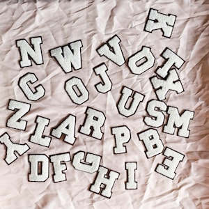 Peut inclure: Une collection d'écussons en forme de lettres de style universitaire, blancs avec des bordures marron foncé, dispersés dans le cadre. Chaque écusson présente une surface texturée et duveteuse ainsi qu'une police en lettres capitales.