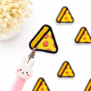 Op de afbeelding: Een driehoekige pizzapunt-patch met gele kaas en rode pepperoni, voorzien van een dikke zwarte rand en een witte omlijning. De patch wordt vastgehouden door een roze en witte clip in de vorm van een konijnenkop.