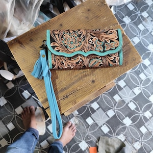 May include: A rectangular leather wallet featuring an intricate floral tooling pattern in shades of brown and black, accented with a scalloped turquoise leather trim and a matching turquoise leather wrist strap with a long fringe tassel.