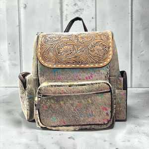 May include: A backpack crafted from gray and white cowhide featuring a vibrant multi-colored paint splatter pattern. The top flap displays intricate floral tooling in tan leather with white stitching. It includes a front pocket and side pockets.