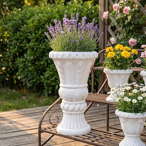May include: A tall, white, pedestal-style planter with ornate relief carvings of roses and leaves, featuring a flared rim and a thick, rope-textured base. The planter is filled with vibrant purple lavender flowers, standing on a wooden deck.