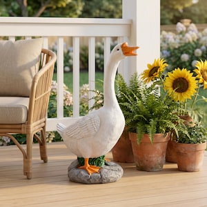 May include: A white goose garden statue with an orange beak and orange webbed feet, standing on a gray textured rock base. The goose is depicted with its neck extended upward and its beak open, featuring detailed feather textures on its body.