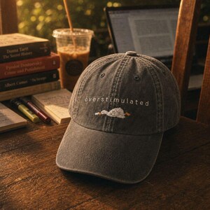 May include: A charcoal grey baseball cap featuring an embroidered white goose lying down with the text "overstimulated" stitched in white letters above the graphic. The cap has a six-panel construction with a curved brim and a button on top.