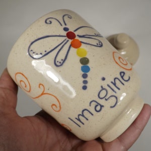 Puede incluir: Taza de cerámica color crema con un diseño de libélula. La libélula tiene alas azules y un cuerpo de puntos de arcoíris. La palabra "imagine" está escrita en azul. Remolinos naranjas decoran el lateral.