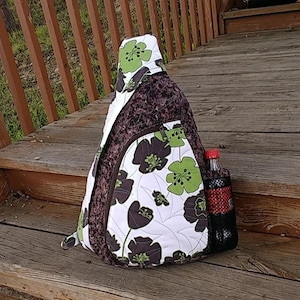 May include: A brown and white floral sling bag with a black mesh side pocket. The bag has a green and brown floral pattern and a brown zipper.