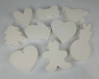 May include: A collection of off-white ceramic ornaments in various shapes, including hearts, a bone, a bird, a Christmas tree, a mitten, a snowman, and a gingerbread man. Each ornament has a small hole for hanging.