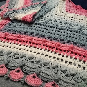 May include: A handmade crochet blanket in shades of pink, white, and gray. The blanket features a textured design with bobbles and a scalloped edge with pink accents. The soft, cozy blanket is perfect for a baby or as a decorative throw.