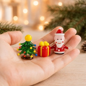 May include: Three miniature glass figurines consisting of a green Christmas tree with colourful ornaments and a yellow star topper, a red gift box with a yellow ribbon, and a Santa Claus figure wearing a red suit and hat with a white beard.