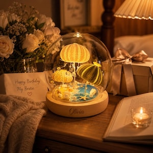 May include: A glass dome display on a round wooden base with the name Olivia engraved on the front. Inside the dome are three sea urchin shells illuminated by small lights, arranged on a blue and sandy base with small seashells and a starfish.