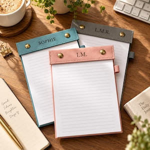 Puede incluir: Tres blocs de notas rayados con cabezales de cuero sintético en azul verdoso, rosa y gris. Cada cabezal tiene dos remaches metálicos de color latón y está personalizado con texto: SOPHIE en el azul, I.M. en el rosa y L.M.R. en el gris.