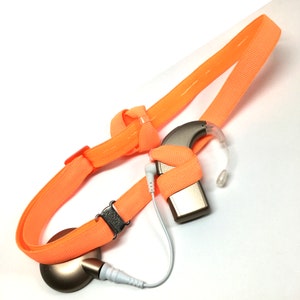 以下が含まれることがあります： オレンジ色の補聴器ストラップに補聴器が取り付けられています。ストラップは柔らかく伸縮性のある素材でできており、バックル留めになっています。補聴器は、マイクとスピーカーが付いた小さなベージュ色のデバイスです。