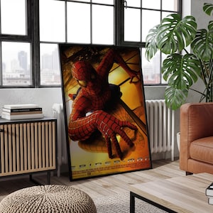 May include: A framed movie poster featuring Spider-Man in a red and blue suit with a black web pattern, crouching against a golden-yellow background. The character is positioned centrally with one hand reaching forward. The title Spider-Man is at the bottom.