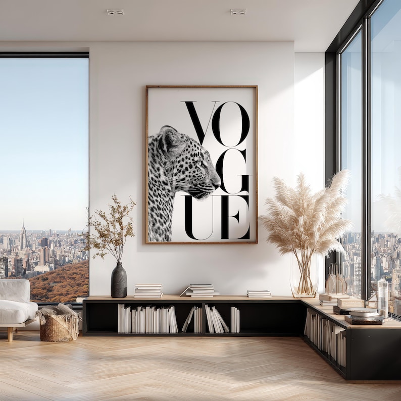 May include: A rectangular wall art print in a wooden frame featuring a black and white profile photograph of a leopard next to the large black text VOGUE. The artwork is displayed on a white wall above a low black shelving unit filled with books.
