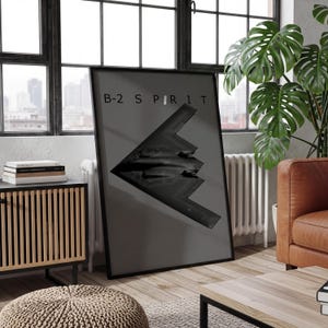 May include: A framed vertical poster featuring a dark gray B-2 Spirit stealth bomber silhouette centered on a solid gray background. The text B-2 S P I R I T is printed in black capital letters across the top portion of the poster.