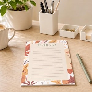 Op de afbeelding: Een rechthoekig notitieblok met een beige middenstuk met de tekst TO-DO LIST en horizontale lijnen, omringd door een rand van herfstbladeren in tinten oranje, rood en bruin. Het notitieblok ligt op een houten oppervlak naast een groene pen.
