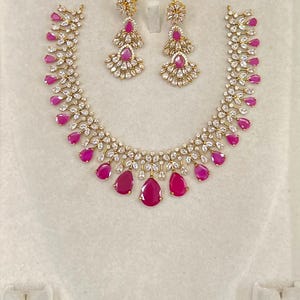 May include: A jewellery set consisting of a necklace and a pair of matching drop earrings. The pieces feature gold-tone metal settings adorned with clear white marquise-cut stones and vibrant fuchsia pink pear-shaped teardrop gemstones.