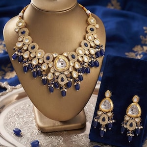 May include: A gold-toned necklace and matching pair of earrings featuring clear, teardrop-shaped stones surrounded by small white crystals and deep blue oval accents. The jewelry set includes dangling blue beads and small white pearls throughout.