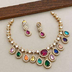 Op de afbeelding: Een goudkleurige ketting en bijpassende oorbellen met veelkleurige, peervormige edelstenen omringd door kleine heldere kristallen. De ketting heeft een centrale ovale rode steen en diverse bungelende druppelvormige edelstenen in roze, groen, oranje, geel, blauw en paars.